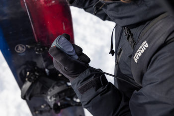 Person adjusting a snowboard with a close-up of their gloved hand, wearing a black jacket with visible branding.