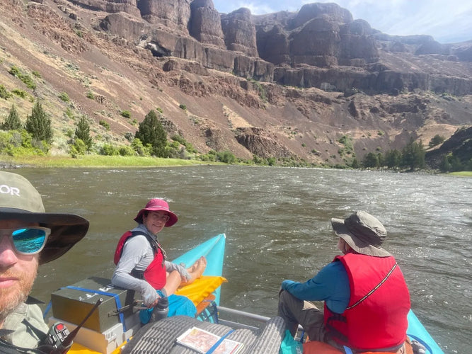Three people in a kayak on a river with mountains in the background