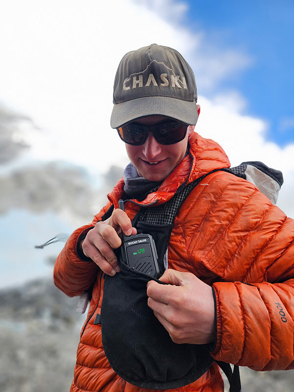 Person in orange jacket and Chaski cap holding a walkie-talkie with mountainous background