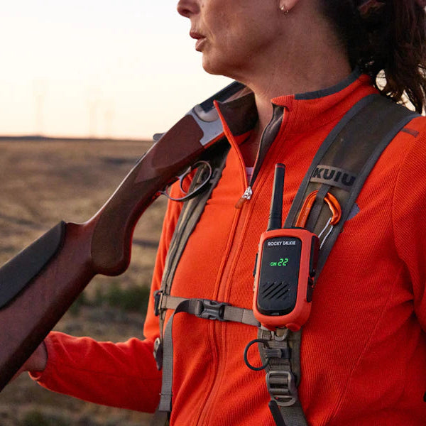 Person in hi-vis orange jacket holding a hunting rifle with an Expedition Radio attached to a strap on her shoulder using a carabiner