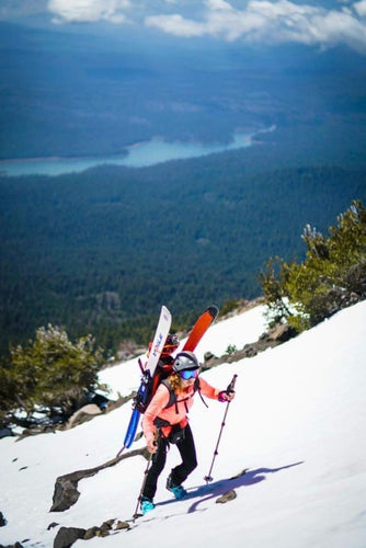 Person with skis and poles hiking on a snowy mountain with a scenic view of a lake and forest.