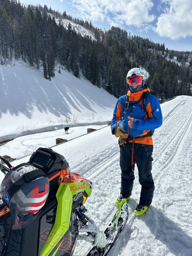 Person in ski gear standing on a snowy slope with a snowmobile nearby