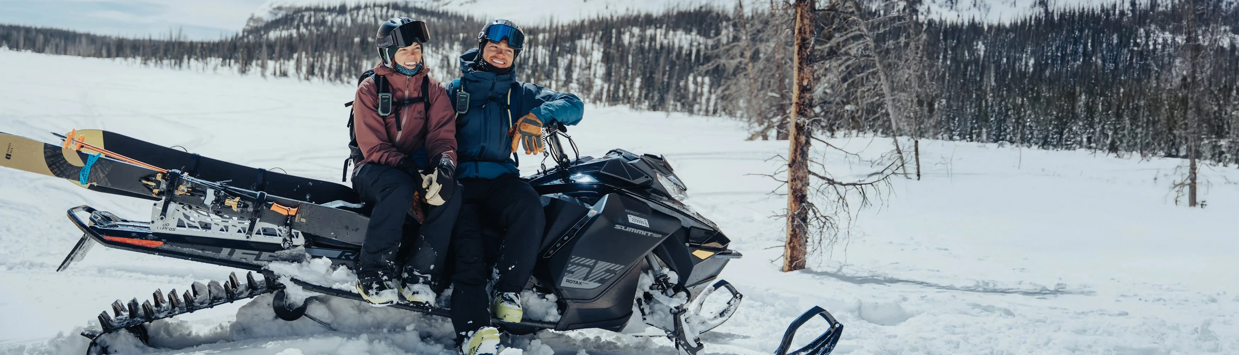 Two people with a snowmobile in a snowy landscape with mountains in the background