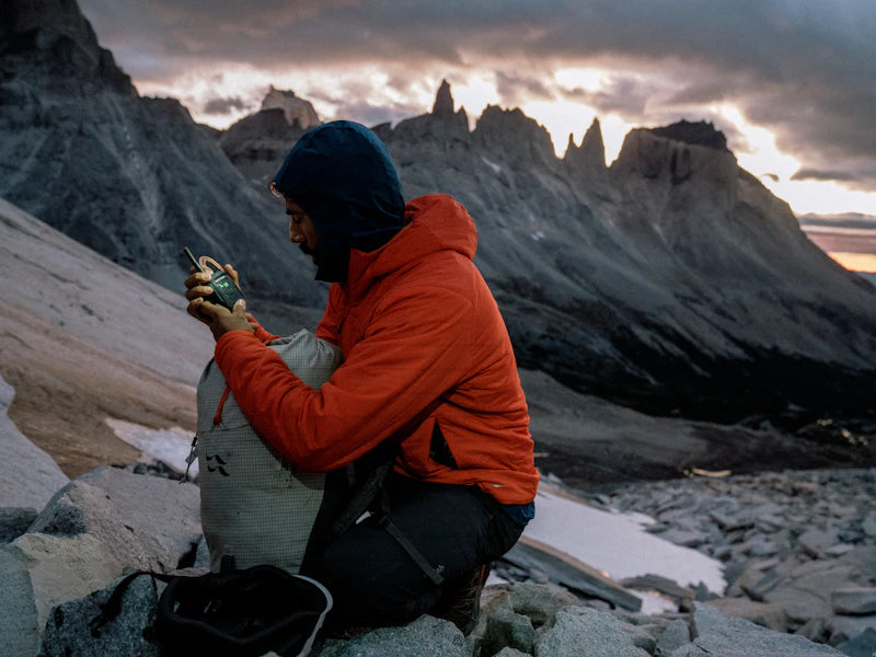 Person in orange jacket sitting on a mountain with snow-capped peaks and dramatic sky.