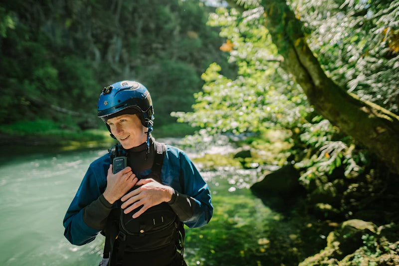 Person wearing a helmet and blue jacket standing by a river with greenery in the background