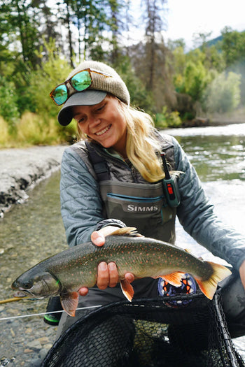 Person holding a fish by a river with a 'Shimms' logo on their clothing
