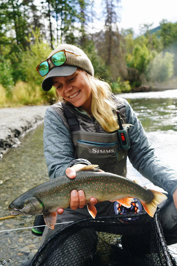 Person holding a fish by a river with a 'Shimms' logo on their clothing