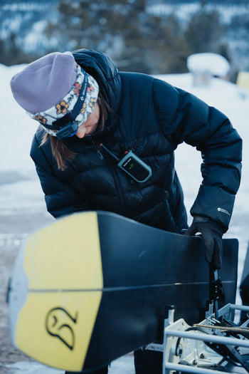 person attaching split board to snowmobile rack in snow with radio on chest