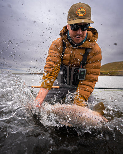 Man in a brown jacket and cap holding a fish caught in water, with a cloudy sky.