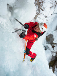 Person ice climbing in a red jacket and helmet on a snowy mountain.
