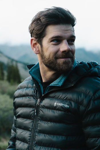 Man wearing a black puffer jacket with a brand logo, standing outdoors with mountains in the background.
