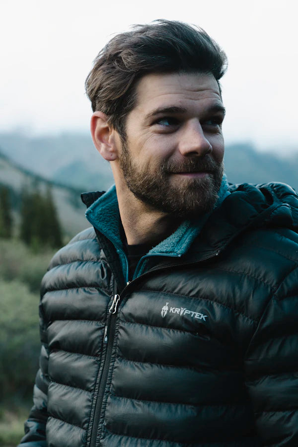 Man wearing a black puffer jacket with a brand logo, standing outdoors with mountains in the background.