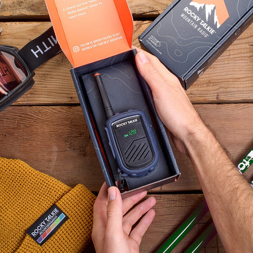 Person opening a box containing a radio on a wooden surface with outdoor gear around