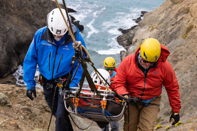 Two rescuers carrying a stretcher with a person on it along a rocky coastline.