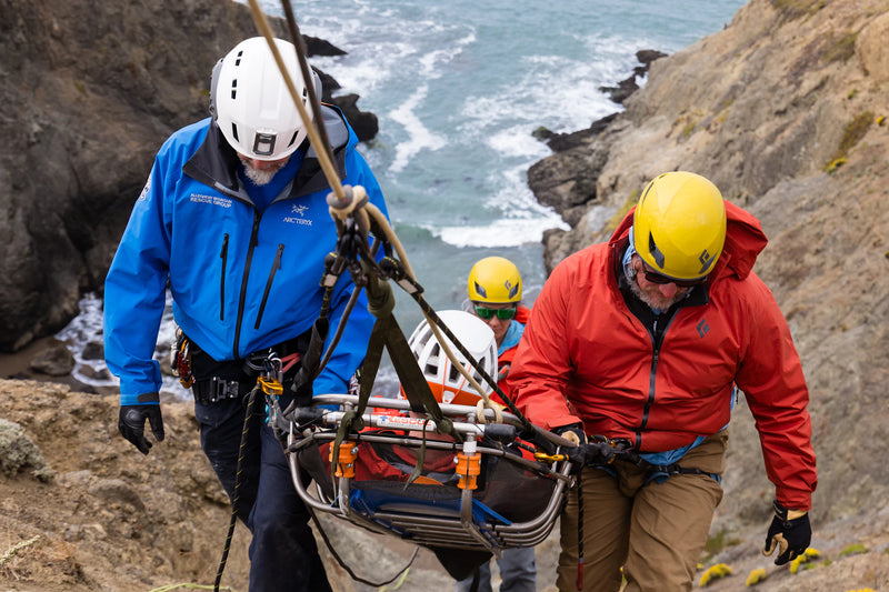 Two rescuers carrying a stretcher with a person on it along a rocky coastline.