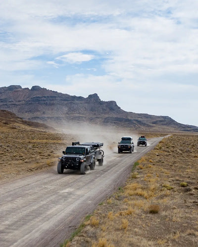 Three off-road vehicles driving on a dirt road with desert landscape and mountains in the background.
